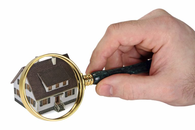 House Inspection with Magnifying Glass in had over house image. A hand holding a magnifying glass over a small model house.