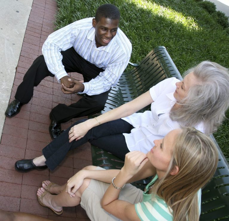 Community Meeting (3 People) Three people conversed while sitting on a bench in a green outdoor setting.