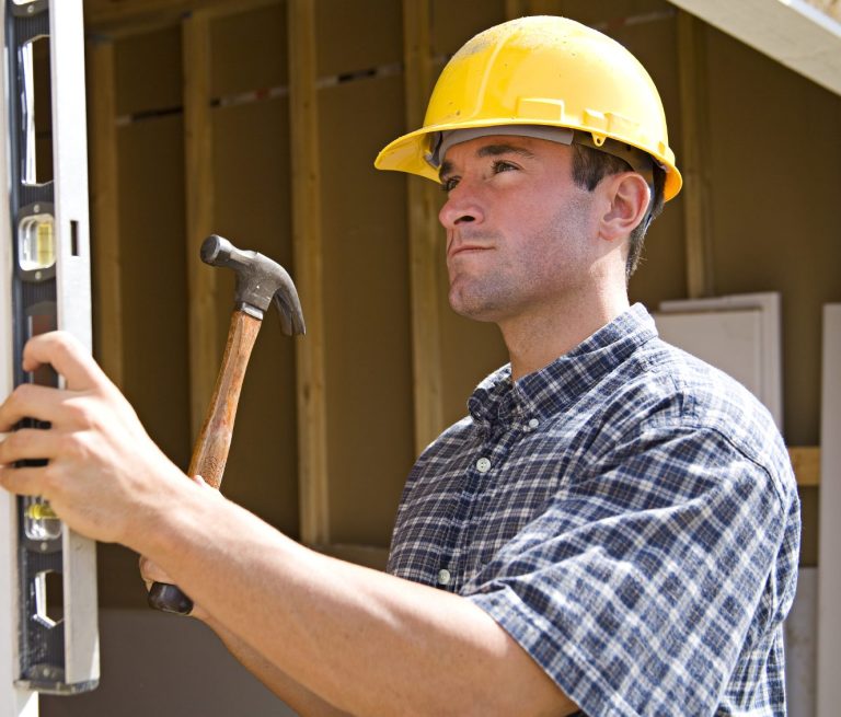 Male Contractor, Leveling and Hammering Wood A construction worker uses a level, holds a hammer, and wears a hard hat.