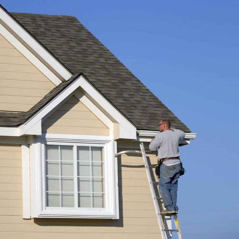 Contractor repairing vinvl house siding A man on a ladder repairing the eaves of a beige house under a clear blue sky.