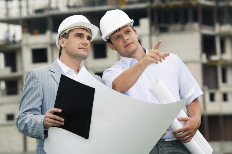 Male Contractor Giving Direction to other Male Contractor Two men in hard hats examine blueprints at a construction site.