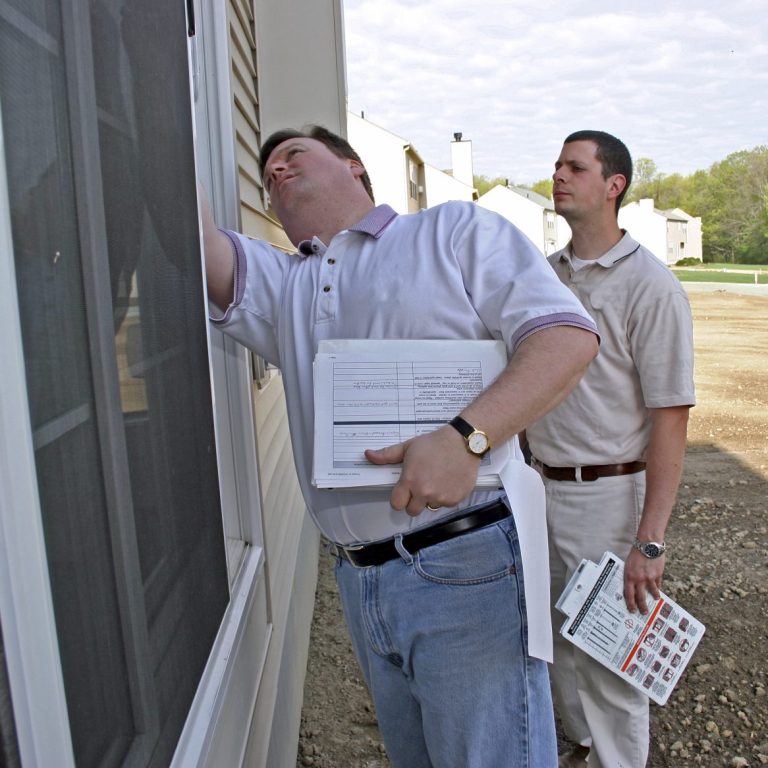 Two Men Inspect Home Two men inspecting a building, one examining a window and the other holding documents.