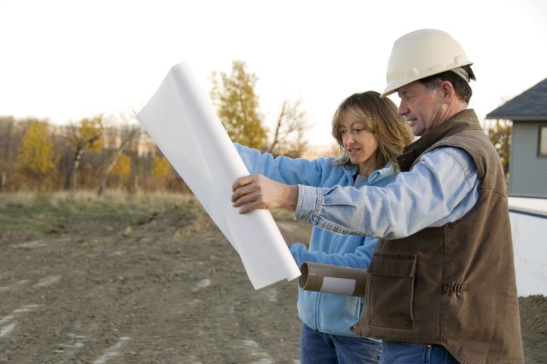 Male Contractor reviews blueprint with female client. Two construction workers review blueprints outdoors at a building site.
