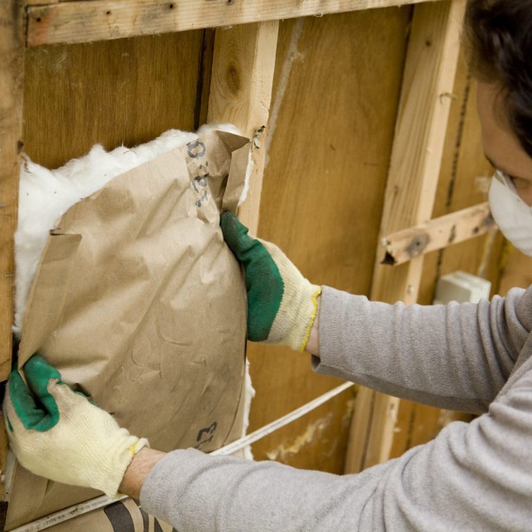 Replacement of Home Insulation between Wooden Studs. A person wearing gloves installing insulation in a wall cavity.