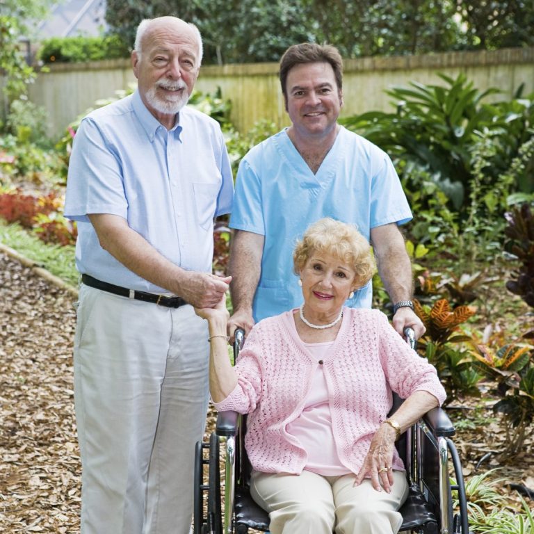 Elderly Couple with Caregiver Two men in a garden surround a smiling elderly woman in a wheelchair.
